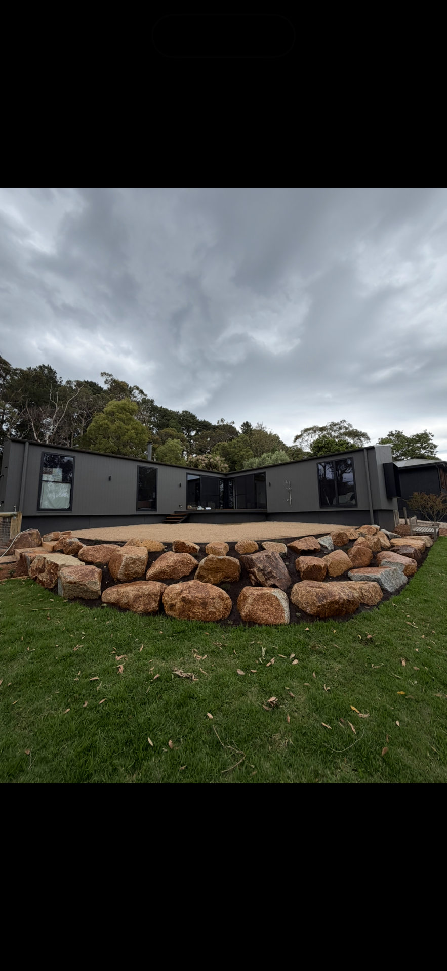 Curved granite boulder retaining wall wrapping a raised gravel terrace behind a modern black-clad home.