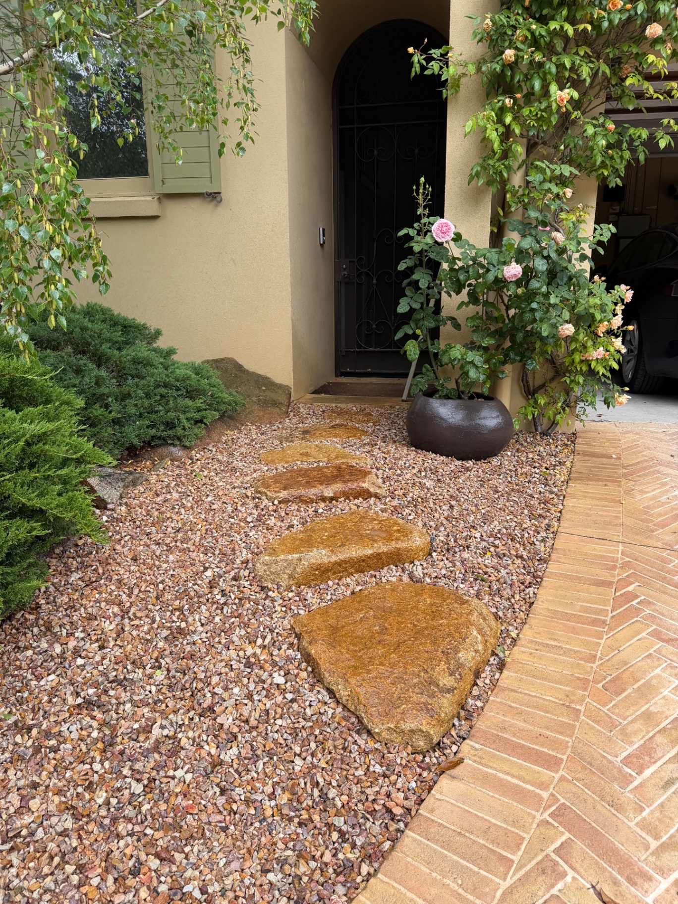 Natural sandstone stepping stones in a warm pebble bed at a rendered home entry, framed by conifers and a climbing rose.