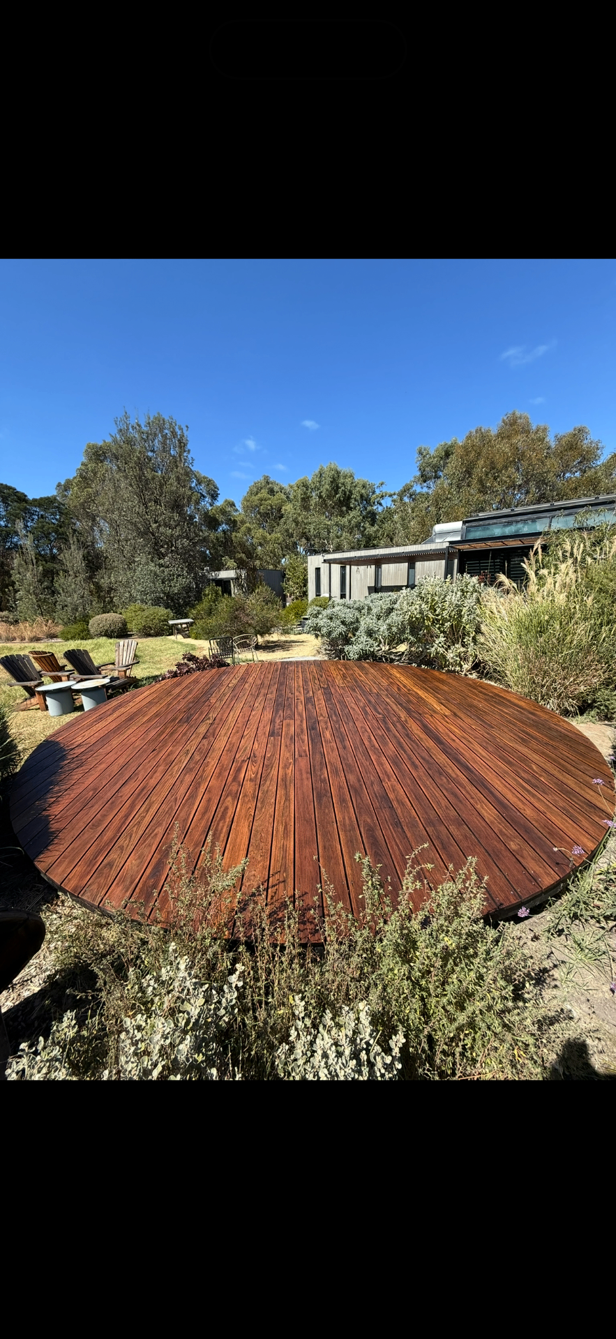 Circular hardwood deck oiled a deep red, set into a native garden with adirondack chairs alongside.